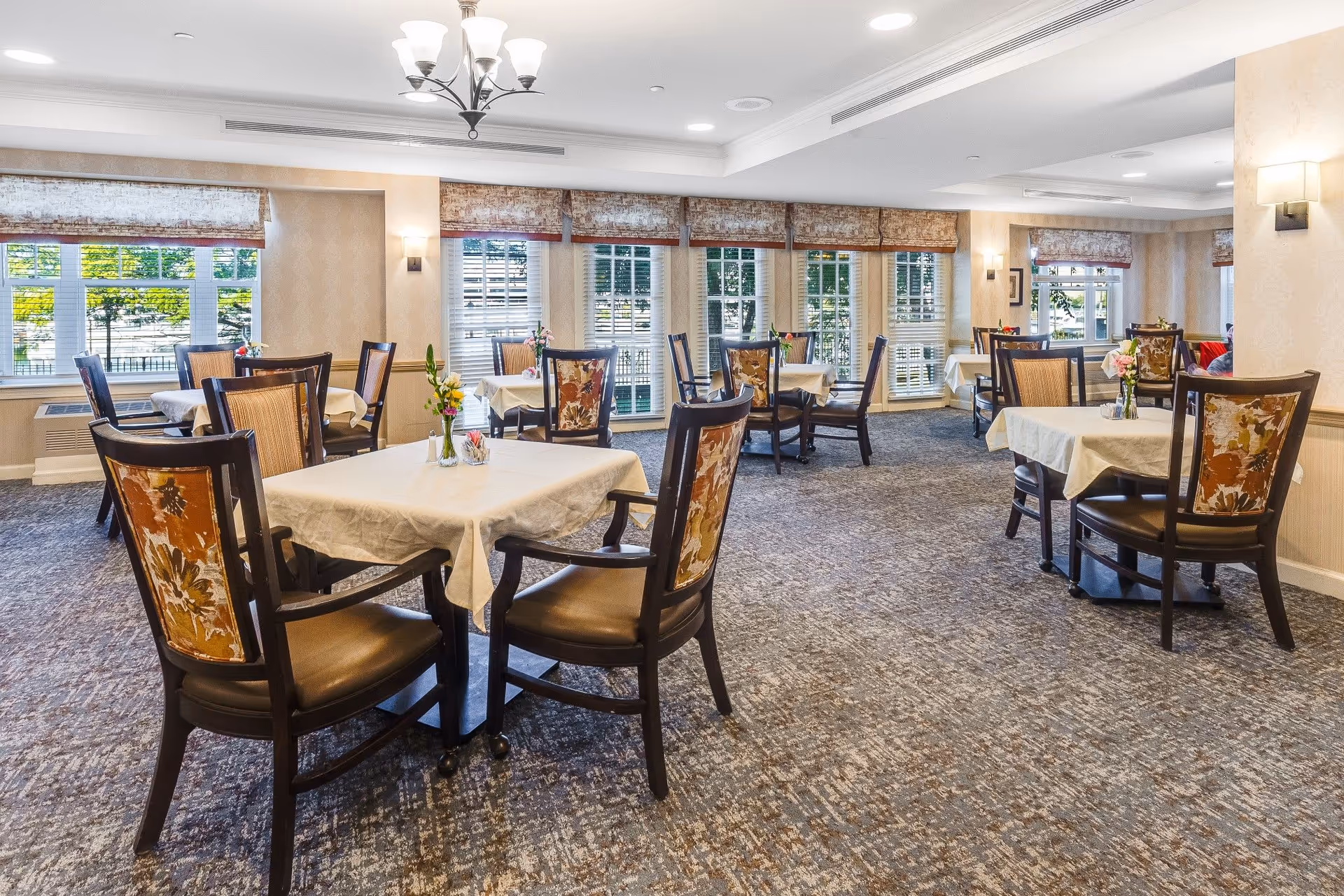 A bright and spacious dining room with multiple tables covered in white tablecloths, each surrounded by wooden chairs with floral upholstery. The room features large windows with blinds and valances, allowing natural light to fill the space. Small flower arrangements are placed on each table, and a chandelier hangs from the ceiling.
