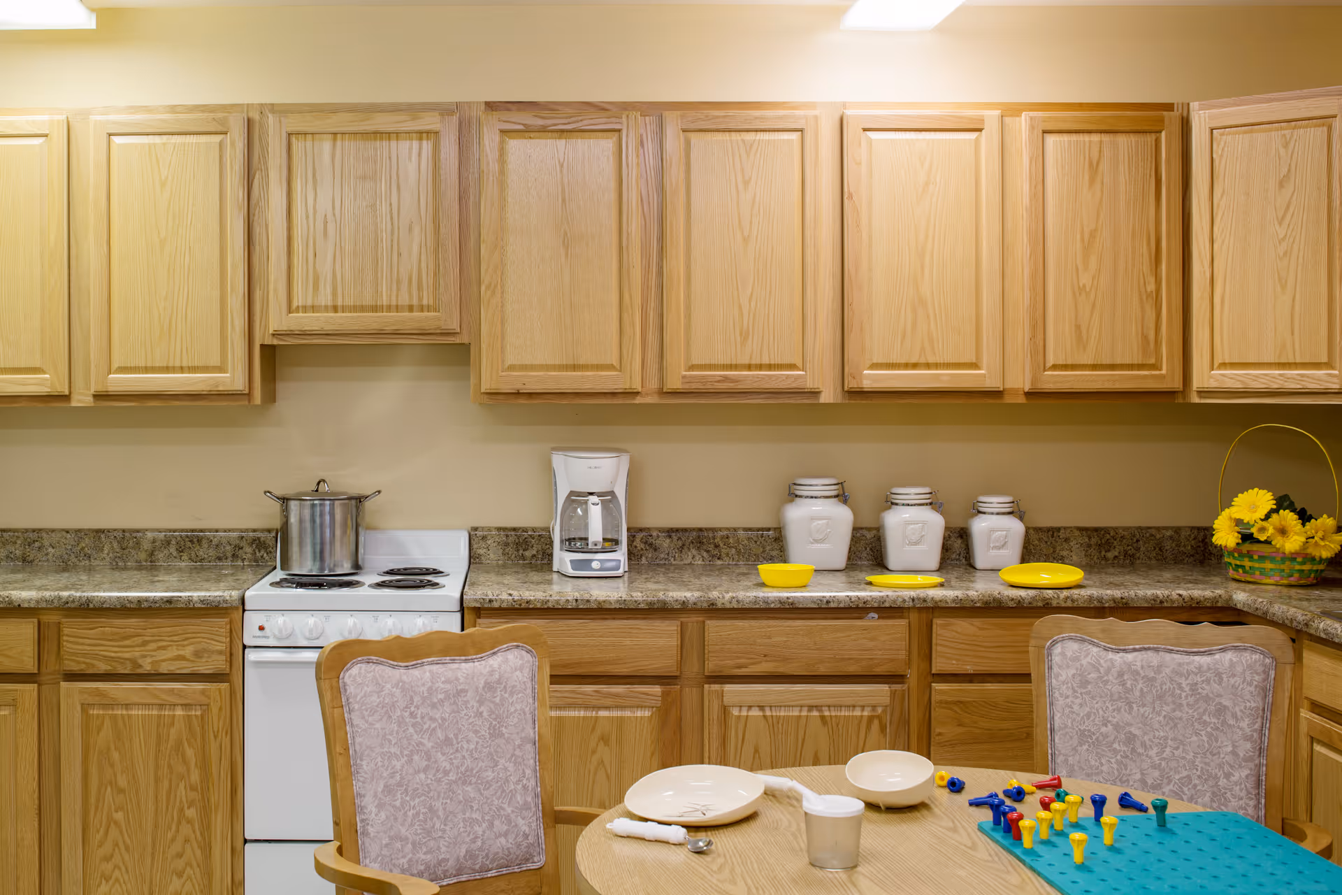 Kitchen with wooden cabinets, a stove and coffee maker on the counter, and a table set with dishes and a pegboard game.