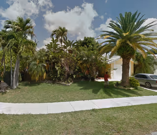 A residential building surrounded by lush palm trees and greenery under a partly cloudy sky. A person in a red shirt is standing near the driveway where a silver car is parked.