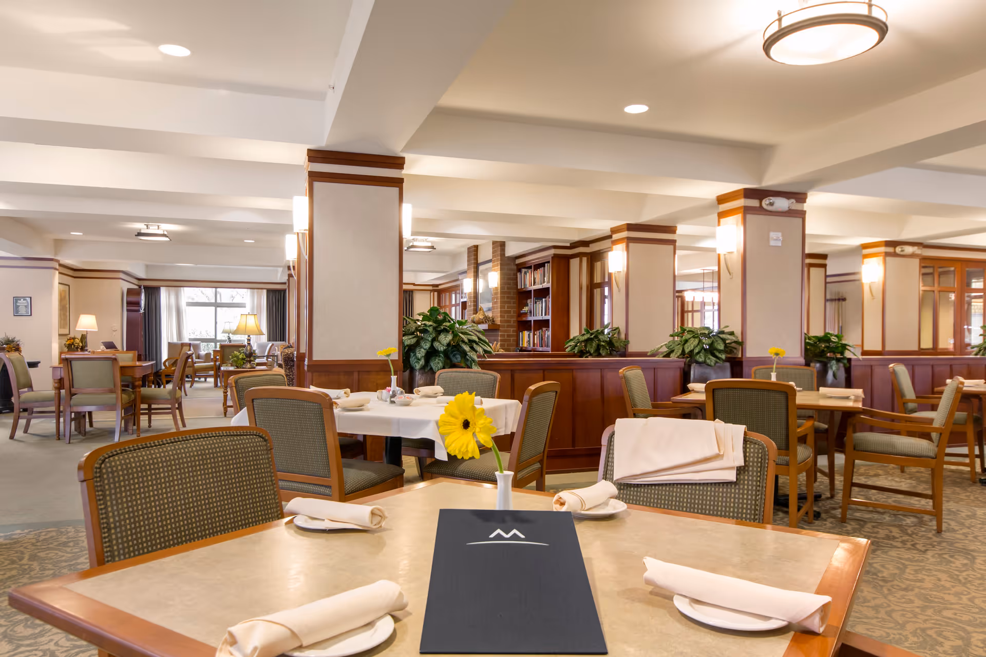 A spacious and well-lit dining room with multiple tables set with white napkins and plates. Each table has a small vase with a yellow flower. The room features wooden paneling, green cushioned chairs, plants, and soft lighting fixtures on the walls and ceiling. Large windows in the background allow natural light to enter the space.