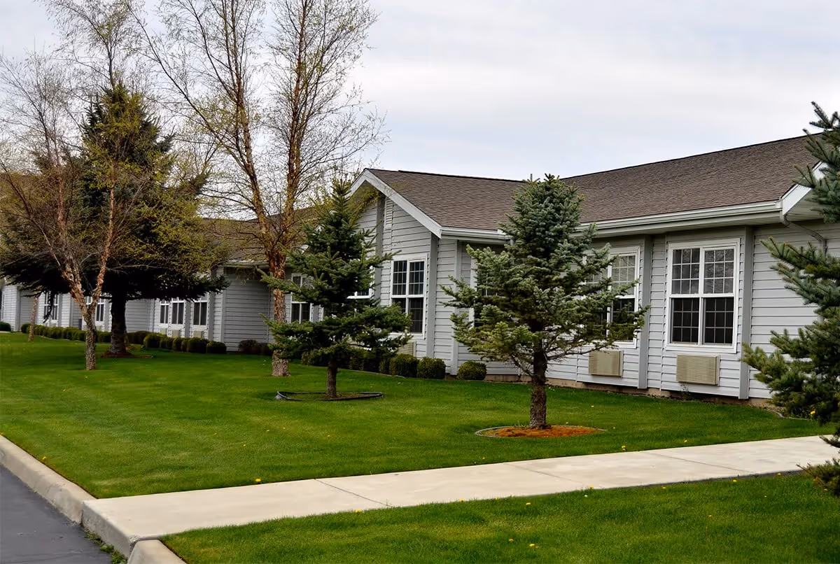 Exterior view of a single-story senior living facility building with gray siding, multiple white-framed windows, and a well-maintained green lawn with small trees and a sidewalk in front.