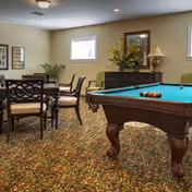 A cozy interior room featuring a pool table with billiard balls arranged on it, a wooden dining table with chairs, a sideboard with a lamp and floral arrangement, and framed artwork on the walls. The room has a patterned carpet and a small window letting in natural light.