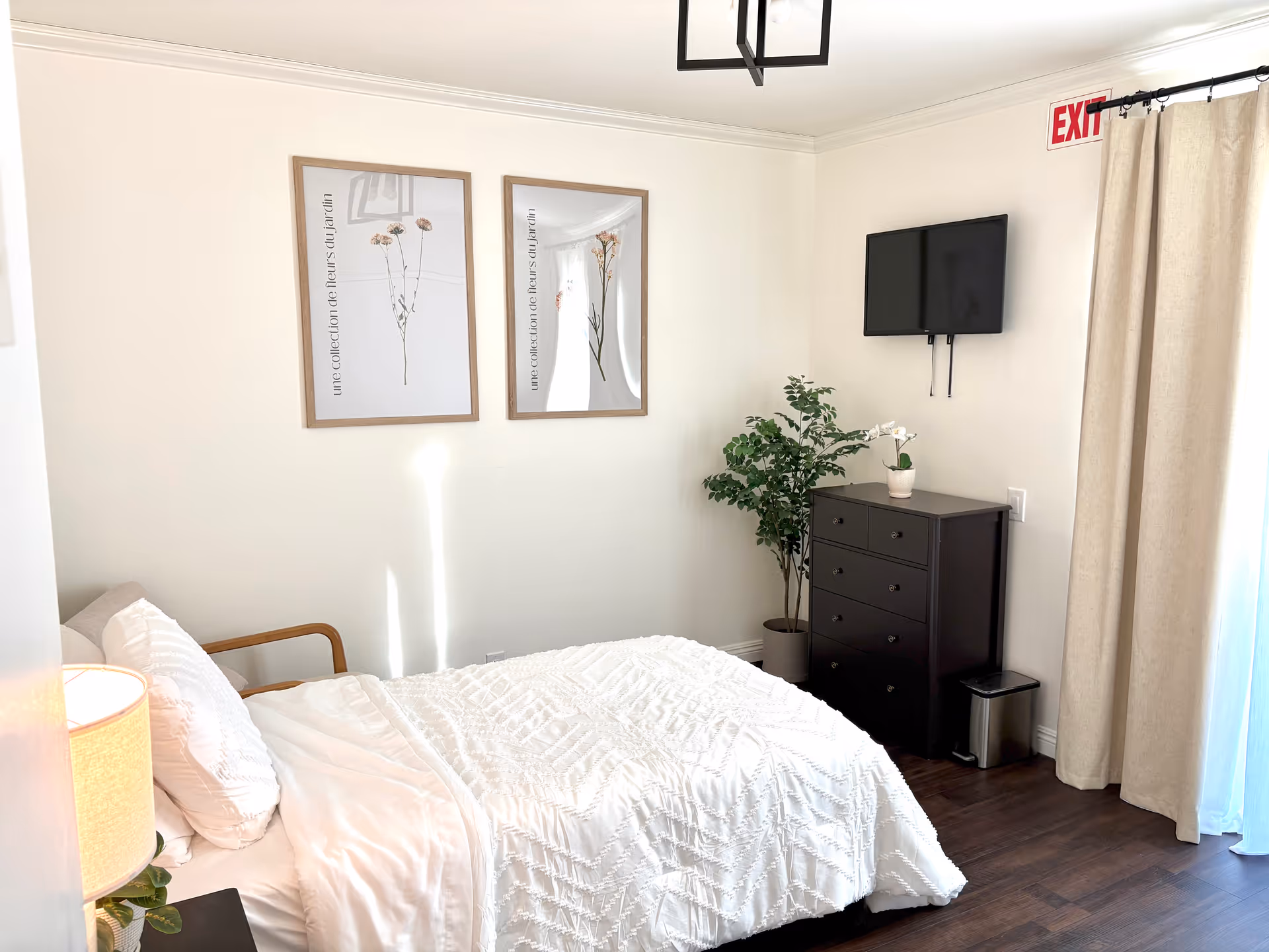 A bright bedroom with a white bedspread on a bed, a wooden armchair partially visible, a bedside lamp, two framed floral prints on the wall, a black dresser with a small potted plant on top, a wall-mounted TV, a green potted plant beside the dresser, beige curtains covering a window or door, and an exit sign above the curtains.