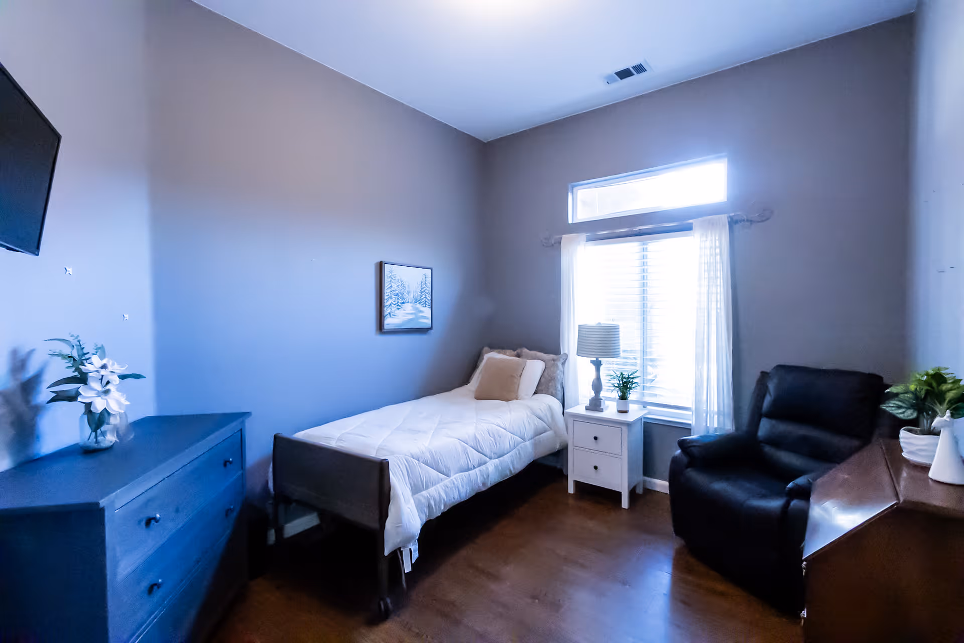 A small, cozy bedroom with a single bed covered in a white comforter and beige pillows. Next to the bed is a white nightstand with a lamp and a small plant. There is a black recliner chair near the window, which has white curtains and a transom window above it. A dark blue dresser with a vase of flowers is on the left side of the room, and a flat-screen TV is mounted on the wall above it. The walls are painted light gray and the floor is wooden.