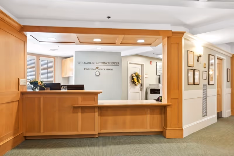Reception desk area in The Gables at Winchester senior living facility with wooden paneling, a small flower arrangement on the desk, and a wall sign displaying the facility name and Five Star Senior Living logo. The hallway to the right has framed pictures and a door decorated with a wreath.