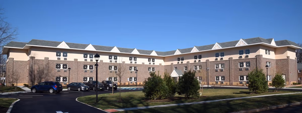 Exterior view of a large, three-story senior living facility building with brick and beige walls, multiple windows, a parking lot with several cars, and a well-maintained lawn with small trees under a clear blue sky.