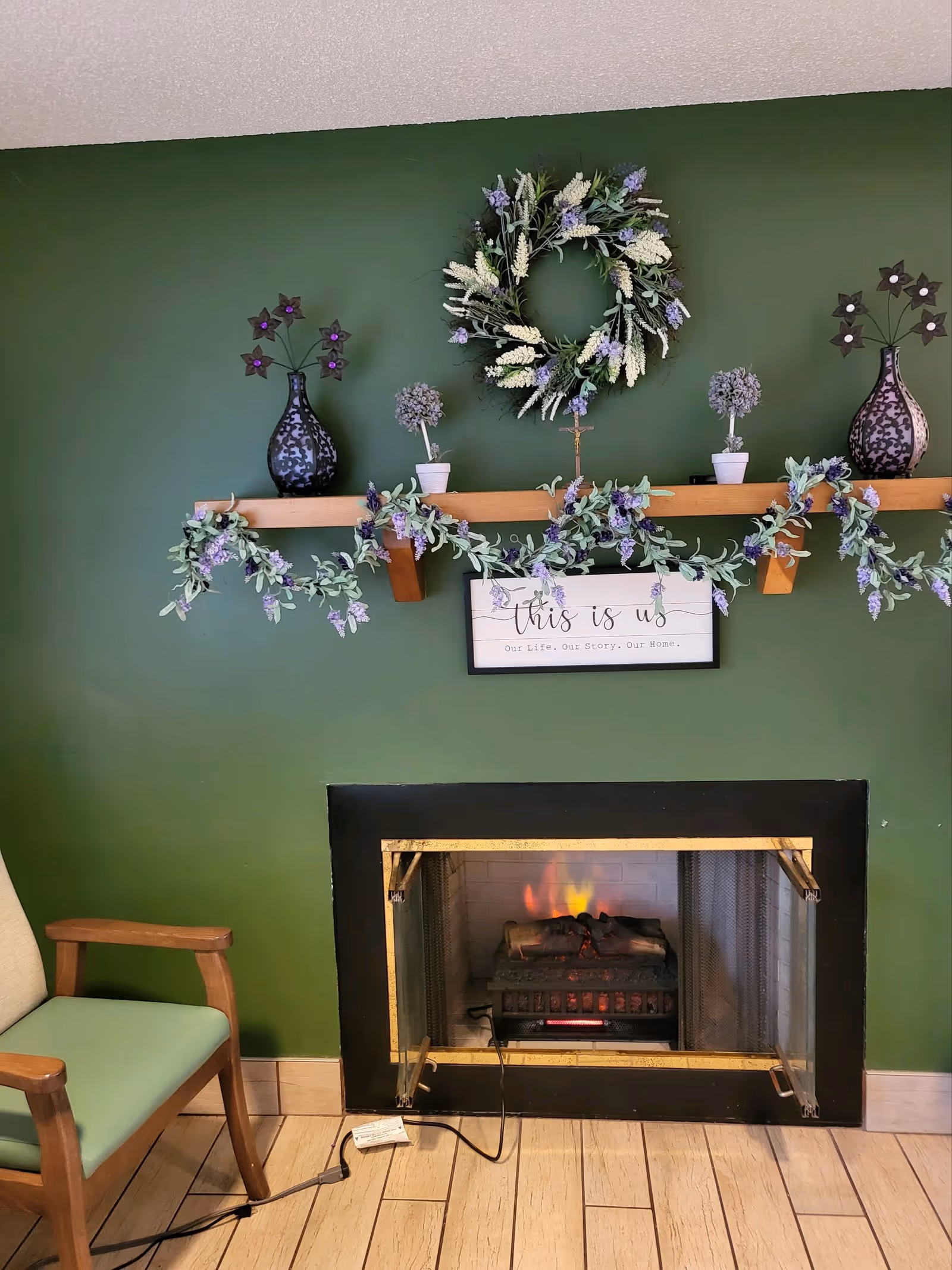 A cozy interior corner featuring a green wall with a wooden shelf decorated with two black vases holding purple flower ornaments, two small potted plants, and a floral wreath. Below the shelf is a framed sign that reads 'this is us Our Life. Our Story. Our Home.' A black-framed fireplace with a glowing electric fire is set into the wall. To the left of the fireplace is a wooden chair with light green cushions. The floor is tiled with light wood-patterned tiles.