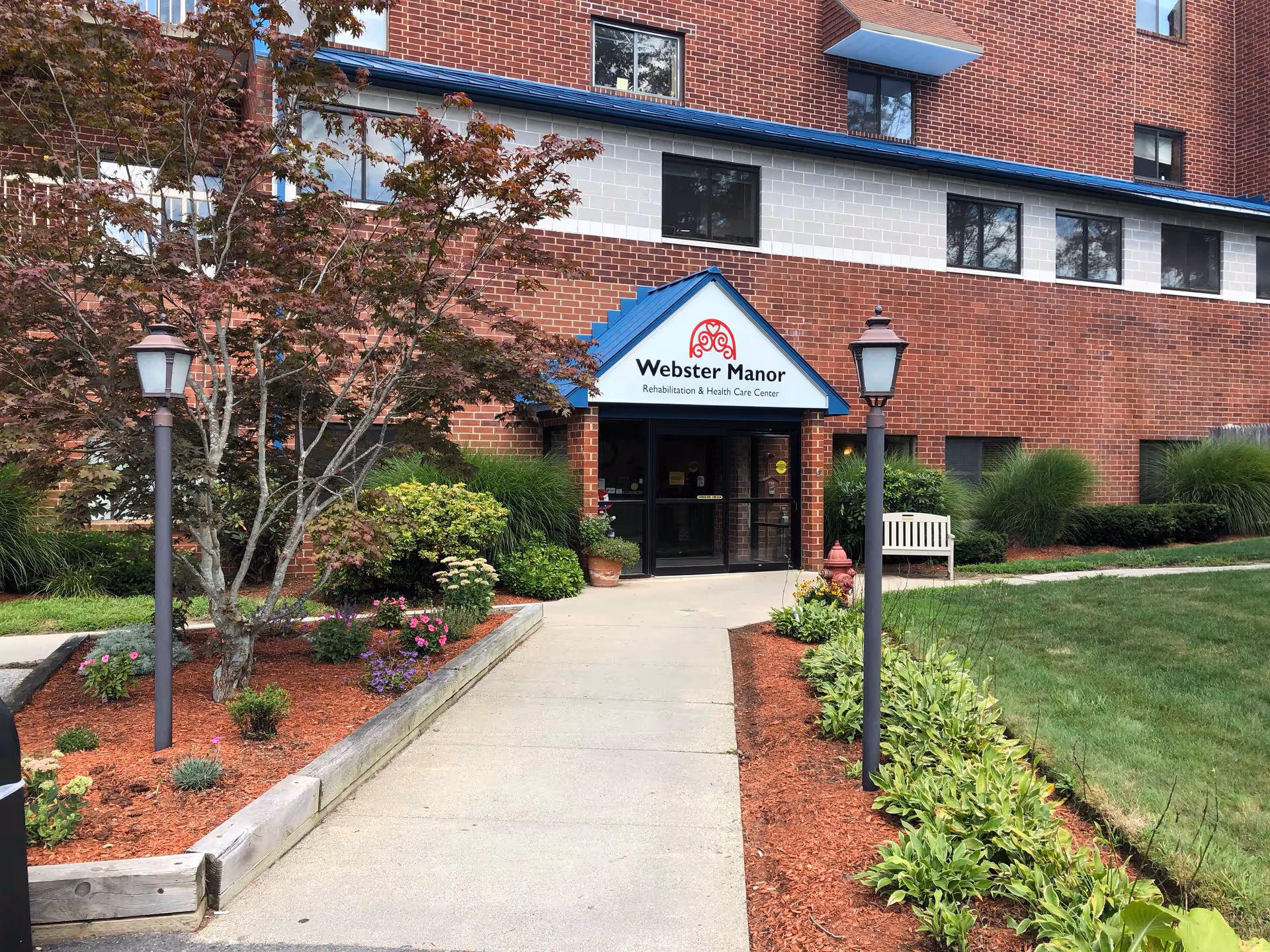 Entrance to Webster Manor Rehabilitation & Health Care Center, a brick building with a blue roof over the doorway, surrounded by landscaped flower beds, bushes, and two lamp posts along the concrete walkway.