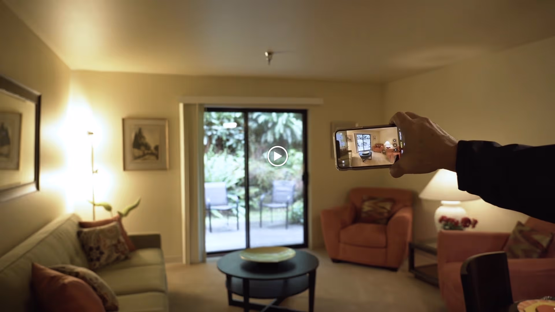 A cozy living room with a green sofa on the left, two orange armchairs on the right, a round black coffee table in the center, and a sliding glass door leading to an outdoor patio with chairs and greenery. A person is holding a smartphone capturing the room in the foreground.