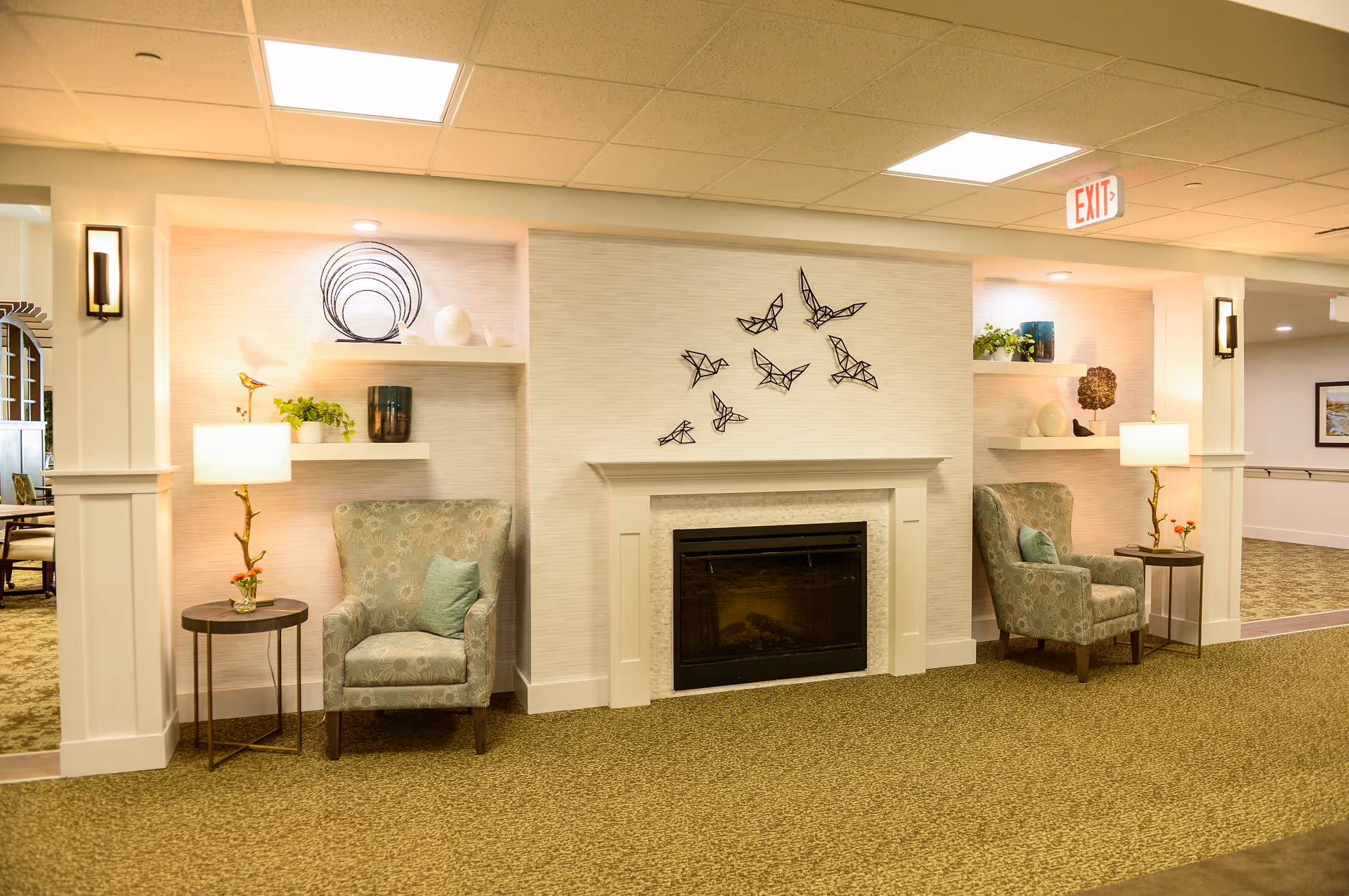 Seating area with a central fireplace flanked by two armchairs, side tables, lamps and decorative shelving in a senior living facility.