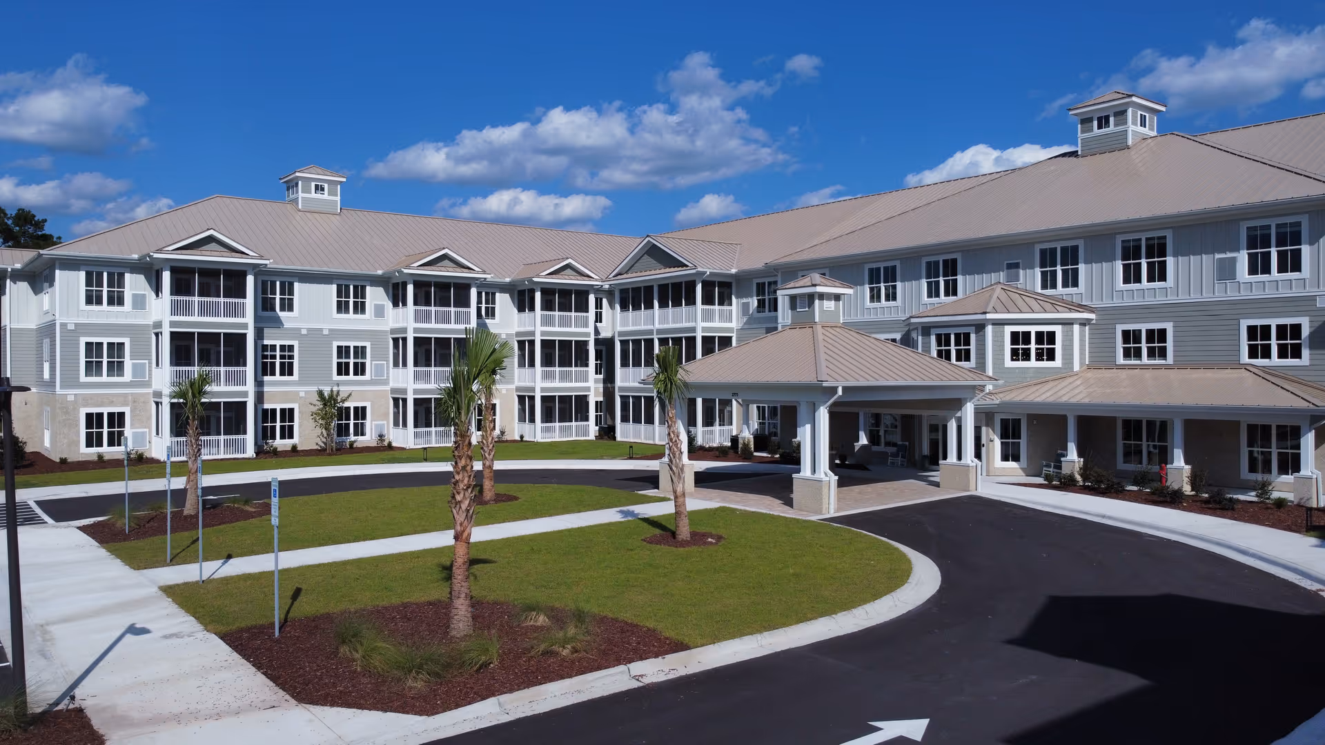 Exterior view of a large, modern senior living facility building with a light gray and white facade, multiple windows, and a covered entrance. The building is surrounded by a circular driveway, green lawn, and several palm trees under a blue sky with scattered clouds.