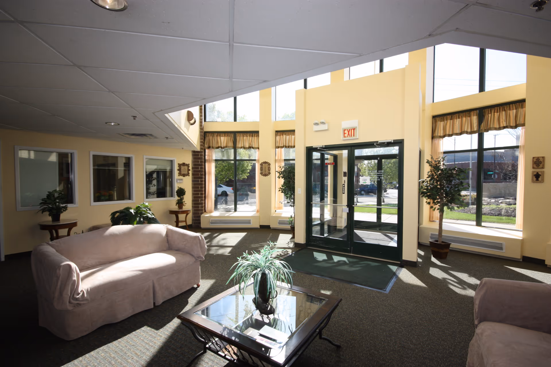 A bright and spacious lobby area with large windows allowing natural light to fill the room. The space features two beige sofas, a glass-top coffee table with a plant centerpiece, potted plants near the windows, and a double glass door exit with an exit sign above it.