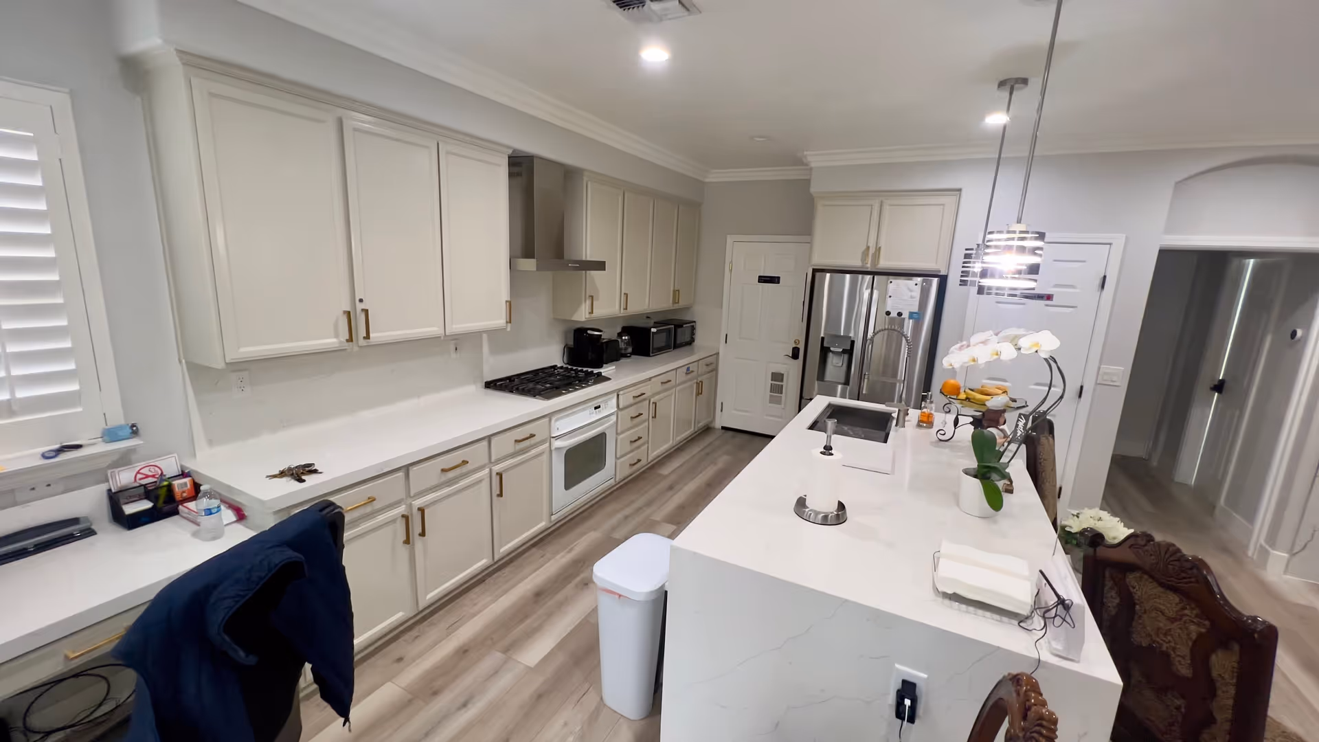 A modern kitchen with white cabinetry and gold handles, a white countertop island with a sink, a stainless steel refrigerator, a stove with a range hood, and various kitchen appliances. There are pendant lights hanging above the island, a trash bin on the floor, and decorative items including a white orchid and a fruit stand on the island. The floor is light wood, and there is a chair with a dark jacket draped over it near a small desk area.