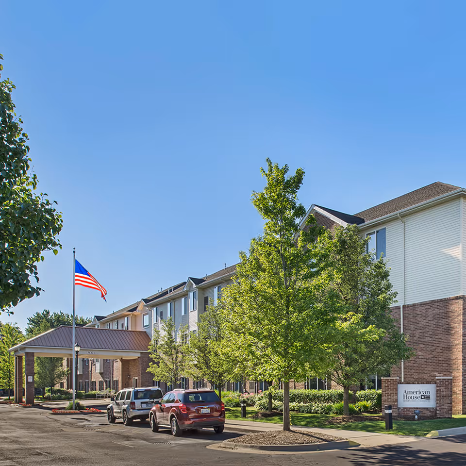 Exterior view of American House Sterling Woods senior living facility on a clear sunny day. The building is three stories tall with a combination of brick and light-colored siding. There is a covered entrance with a metal roof, an American flag on a flagpole, several parked cars, and green trees and landscaping around the property.
