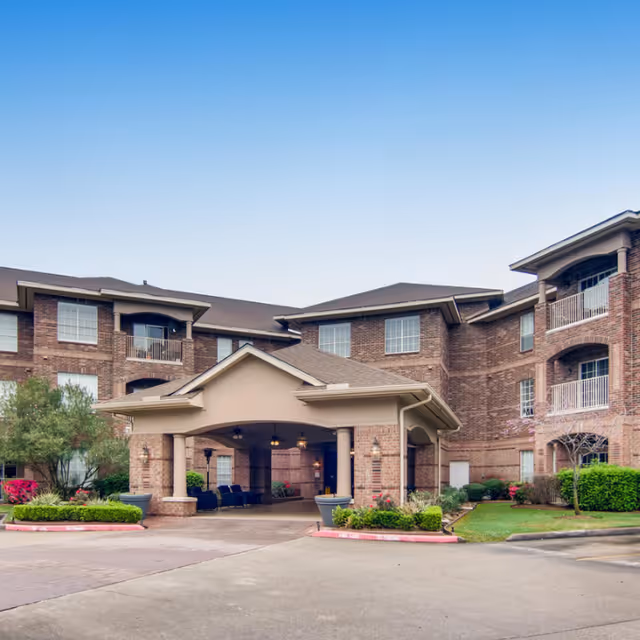 Front entrance of a brick senior living facility with a covered porte-cochère, balconies, and landscaped driveway.