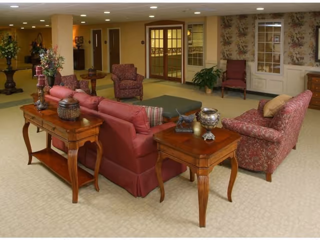 A cozy senior living community lounge area with upholstered armchairs and sofas in red and floral patterns, wooden side tables with decorative items, a green ottoman, floral wallpaper, and a carpeted floor. The background shows double glass doors and a hallway.