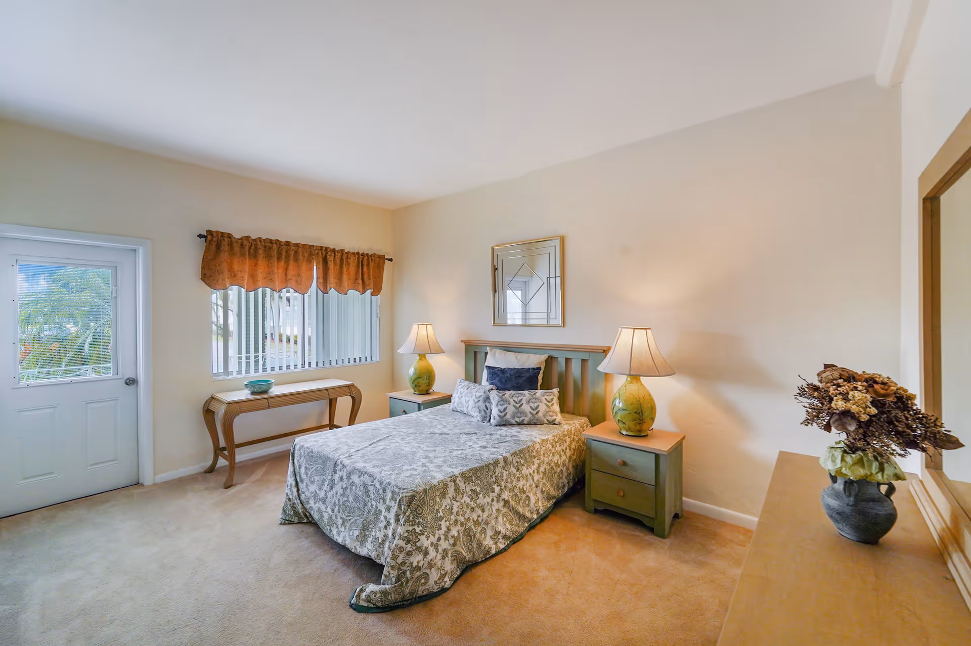 A cozy bedroom with a double bed covered in a patterned bedspread, flanked by two green nightstands each with a lamp. There is a wooden console table under a window with vertical blinds and a door with a window next to it. A large mirror hangs above the bed, and a vase with dried flowers sits on a dresser to the right.