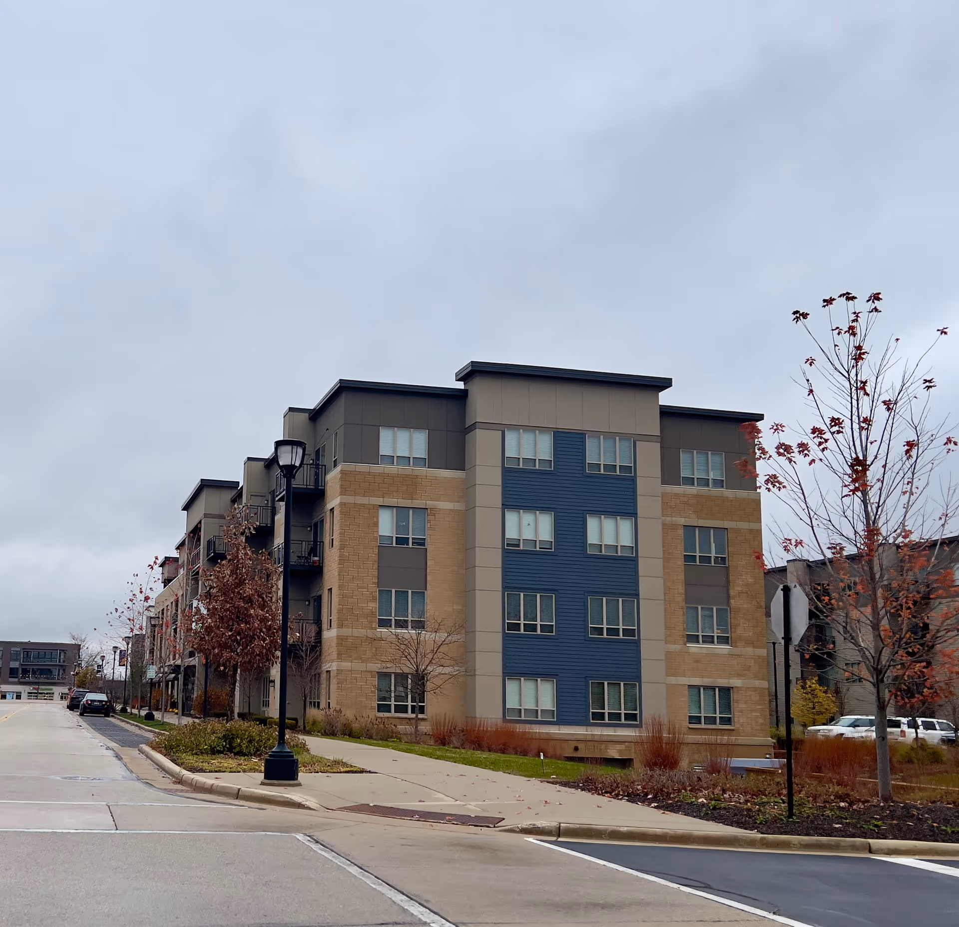 Exterior view of a modern multi-story residential building with beige and blue paneling, surrounded by a sidewalk, street lamps, and some trees with autumn leaves under a cloudy sky.