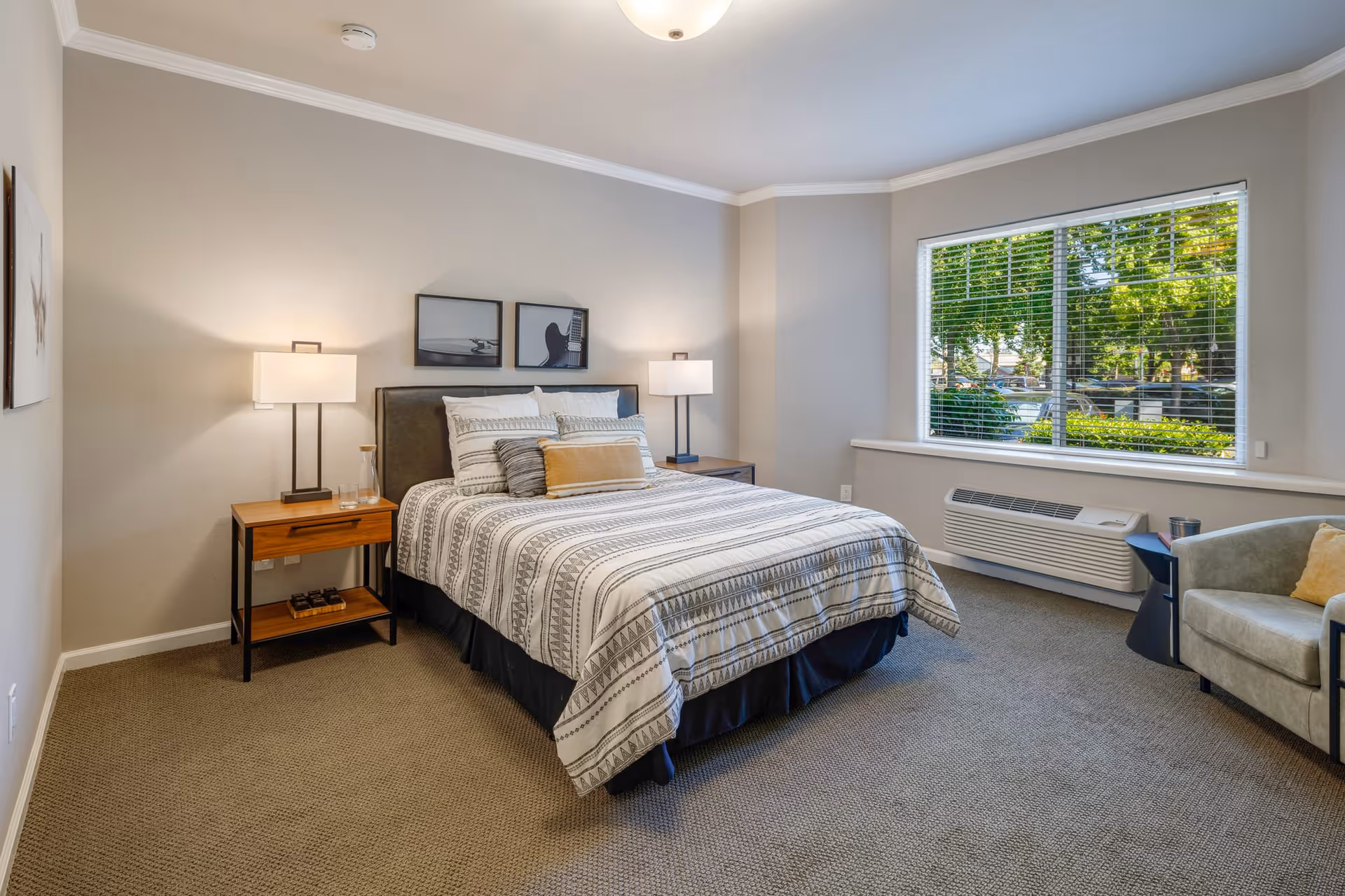 A neatly made bed with patterned bedding in a bedroom featuring two bedside tables with lamps, a large window with blinds showing greenery outside, a beige armchair with a yellow cushion, and neutral-colored walls and carpet.