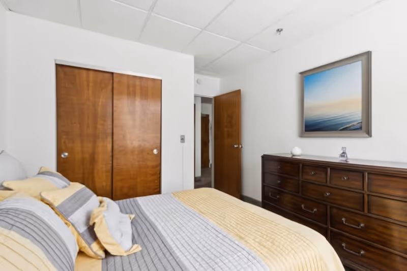A bedroom with a bed covered in gray and yellow bedding, a wooden dresser with multiple drawers, a framed picture of a calm ocean scene on the wall, and wooden closet doors. The room has white walls and a ceiling with recessed lighting.
