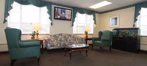Senior living lounge with green chairs, floral sofa, lamps, and a fish tank.