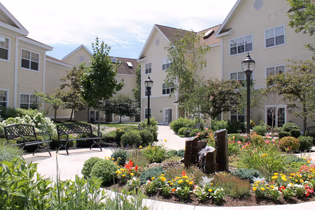 A landscaped outdoor courtyard area with colorful flowers, shrubs, and a small water fountain surrounded by benches and lamp posts. The courtyard is enclosed by a light yellow multi-story building with multiple windows under a partly cloudy sky.