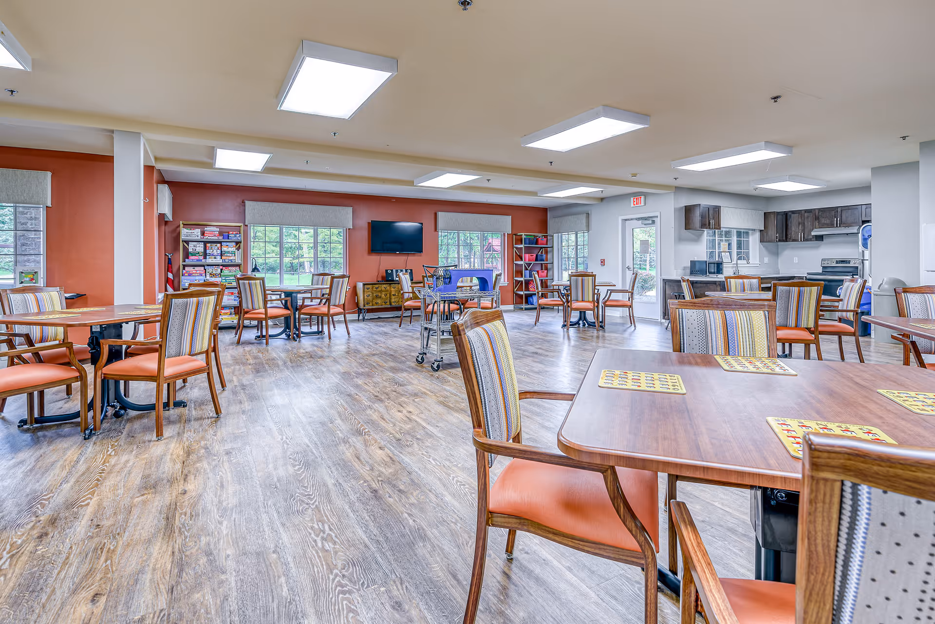 A spacious senior living common area with multiple wooden tables and chairs featuring orange cushions and striped backs. The room has large windows with gray valances, a red accent wall with a mounted flat-screen TV, shelves filled with board games, and a kitchenette area with dark wood cabinets and stainless steel appliances. The floor is wood-patterned, and the ceiling has several rectangular fluorescent lights.