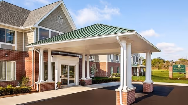Entrance of a senior living facility with a covered drop-off area supported by white columns with brick bases, a green metal roof, and a brick and beige building exterior under a blue sky with some clouds.