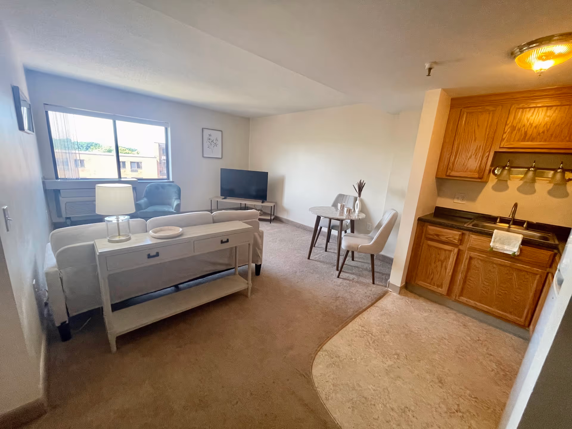 A cozy senior living apartment interior featuring a small living room with a white sofa, a side table with a lamp, a blue armchair near a window, a TV on a stand, and a small round dining table with two chairs. To the right is a compact kitchen area with wooden cabinets, a sink, and overhead lighting.