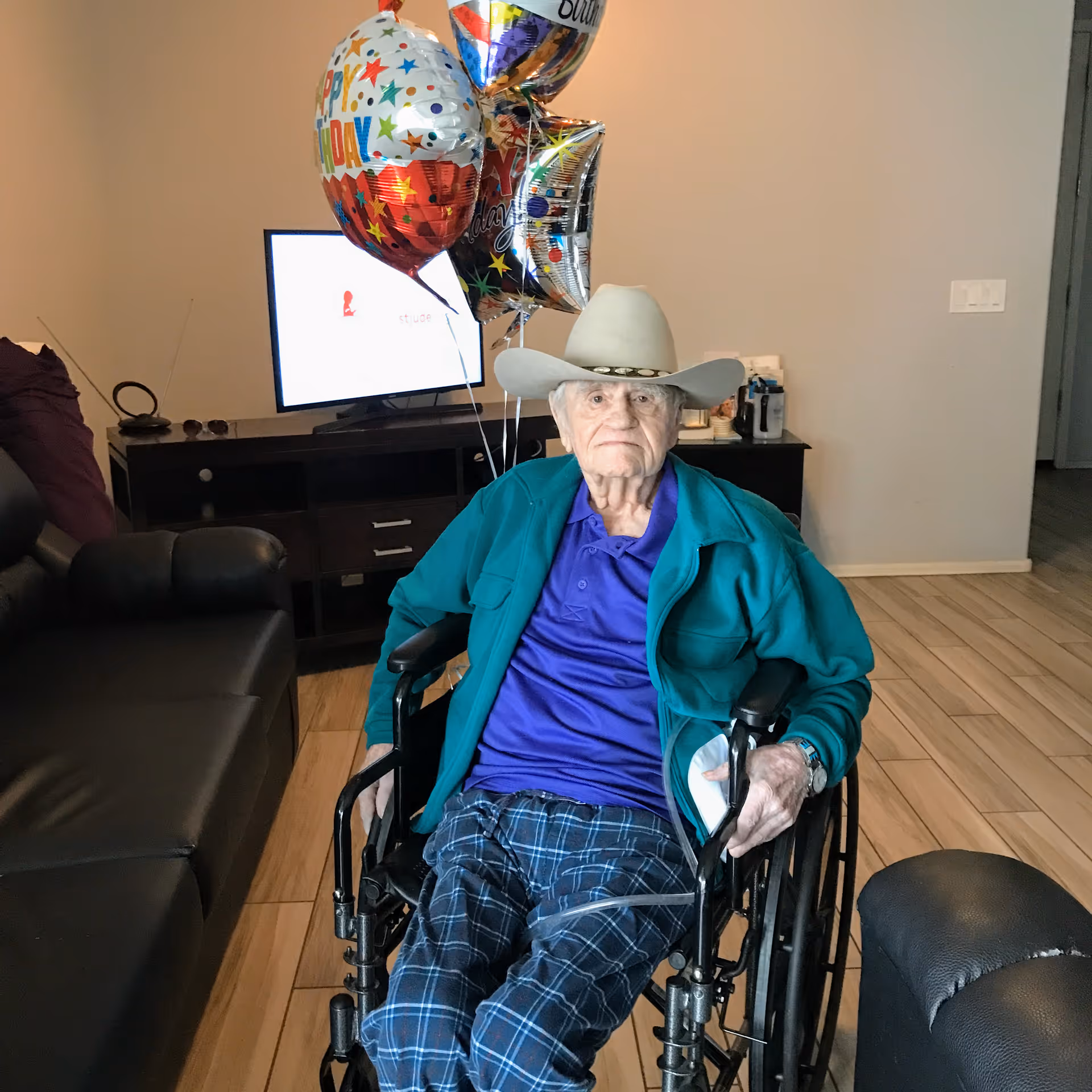 An elderly man wearing a cowboy hat, teal jacket, purple shirt, and plaid pants sits in a wheelchair in a living room. Behind him are colorful birthday balloons and a TV on a dark wooden stand. The room has light-colored wooden flooring and neutral-colored walls.