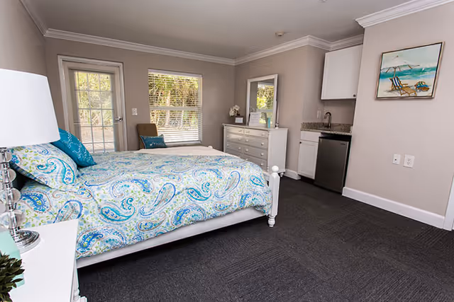 A bright bedroom in Magnolia Manor Assisted Living featuring a bed with a blue and green paisley patterned comforter, a white nightstand with a lamp, a dresser with a mirror, a small kitchenette with a sink and mini fridge, a chair near a window, and a door leading outside.