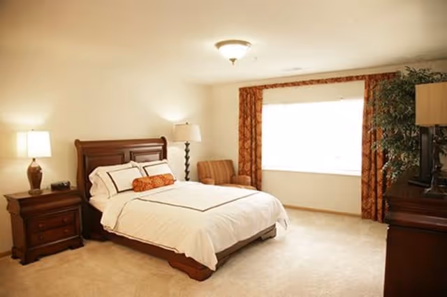 A well-lit bedroom with a large window covered by patterned curtains. The room features a wooden bed with white bedding and decorative pillows, a wooden nightstand with a lamp, an armchair near the window, and a wooden dresser with a TV on top. The carpet is light-colored, and the walls are painted a soft beige.