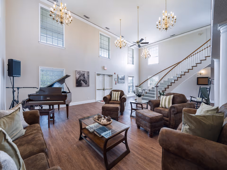 Spacious senior living common area with sofas and armchairs, a grand piano, chandeliers, and a curved staircase.