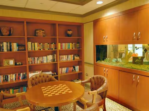 A round table with a chessboard top sits in a library with bookshelves and an aquarium.