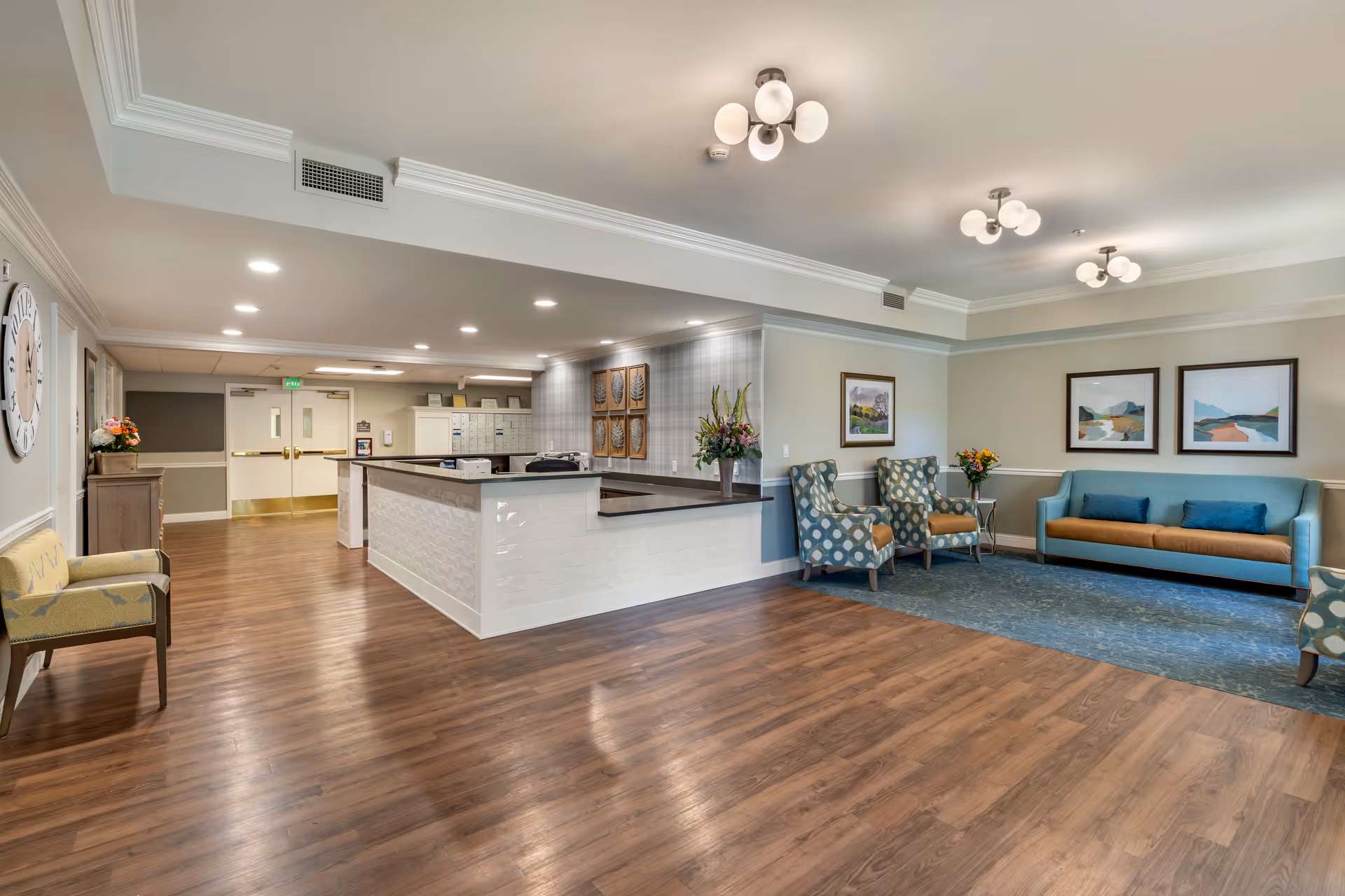Well-lit senior living facility lobby with a reception desk, seating area with sofa and chairs, and wood flooring.