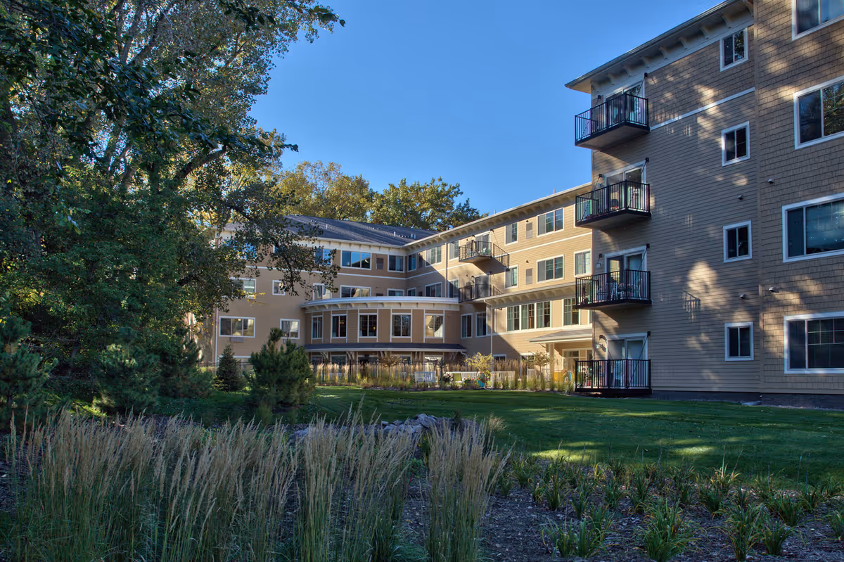 Exterior view of a multi-story senior living facility building with balconies, surrounded by green grass, trees, and landscaping under a clear blue sky.