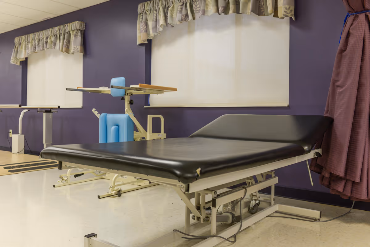 A rehabilitation room with a black padded treatment table, a blue therapy chair with an attached wooden tray, purple walls, and windows covered with white blinds and floral valances.