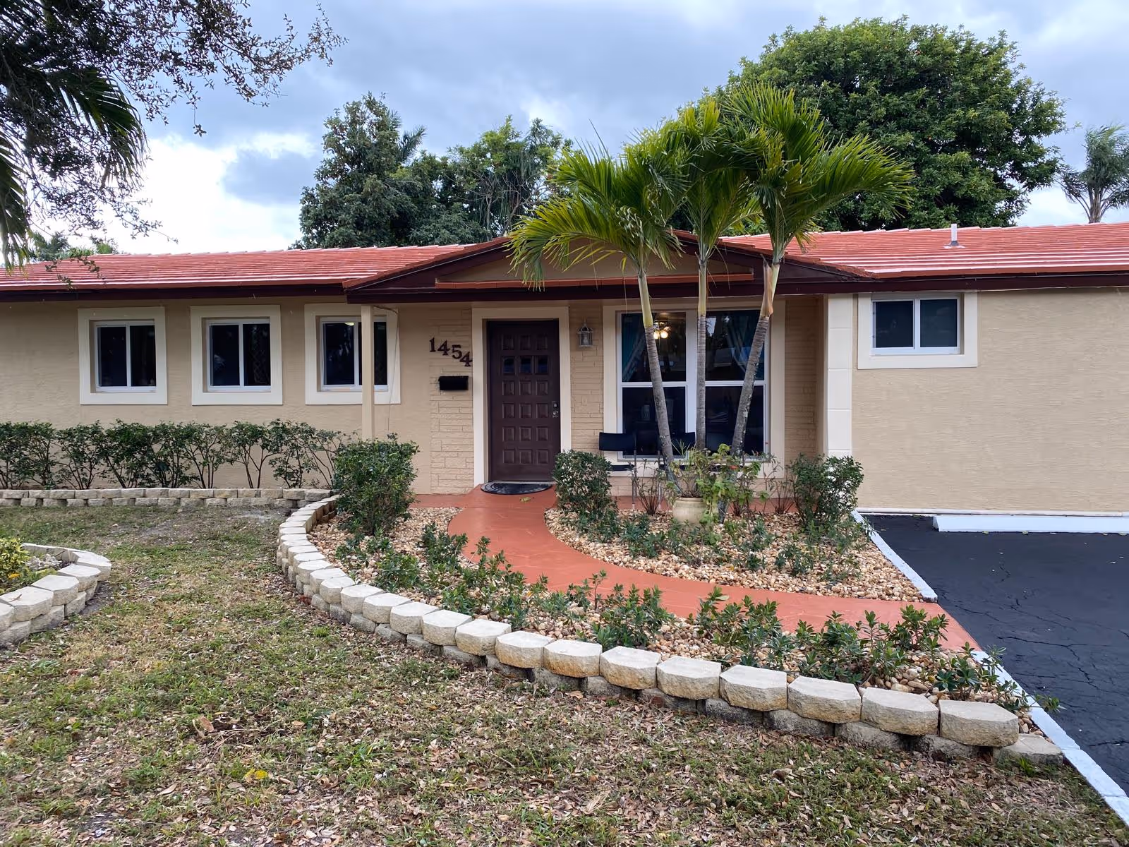Front exterior view of a single-story building with a red roof and beige walls, featuring a brown front door and several windows. There is a curved red walkway leading to the entrance, bordered by small plants and landscaping stones. Two palm trees are planted near the entrance, and there is a driveway visible on the right side. The sky is cloudy.