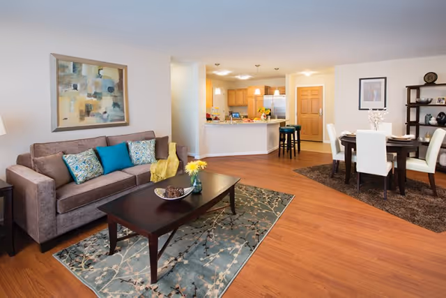 A spacious living and dining area with wooden flooring. The living room features a brown sofa with colorful cushions, a dark wooden coffee table on a patterned rug, and a framed abstract painting on the wall. The dining area has a round dark wooden table with white chairs on a brown rug. In the background, there is a kitchen with wooden cabinets, a refrigerator, and a breakfast bar with two stools.