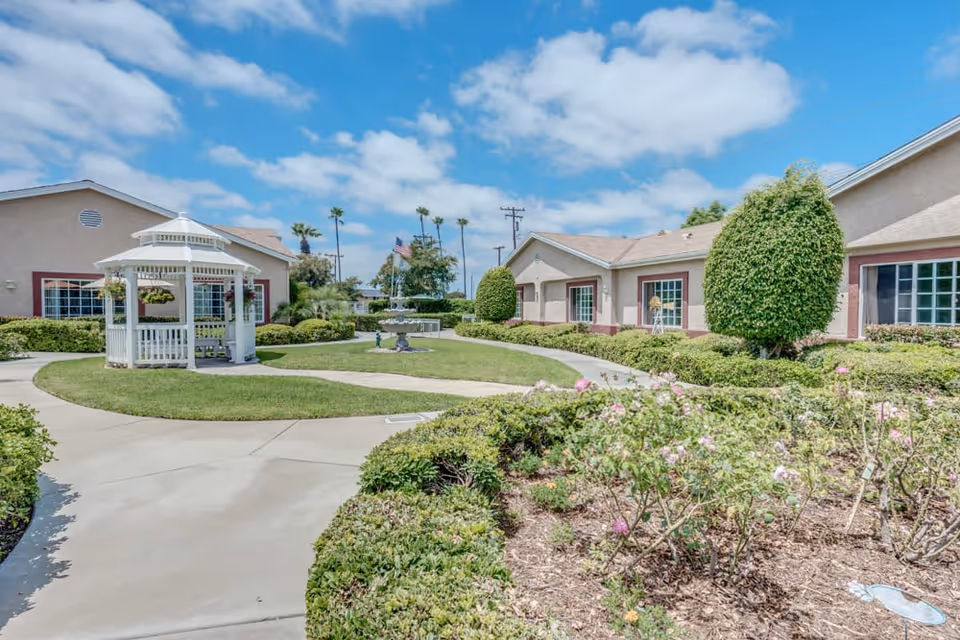 Outdoor garden area at Ivy Terrace at Garden Grove Memory Care featuring a white gazebo, well-maintained shrubs, flowering plants, a fountain, and a pathway leading through the landscaped grounds with single-story buildings in the background under a partly cloudy sky.