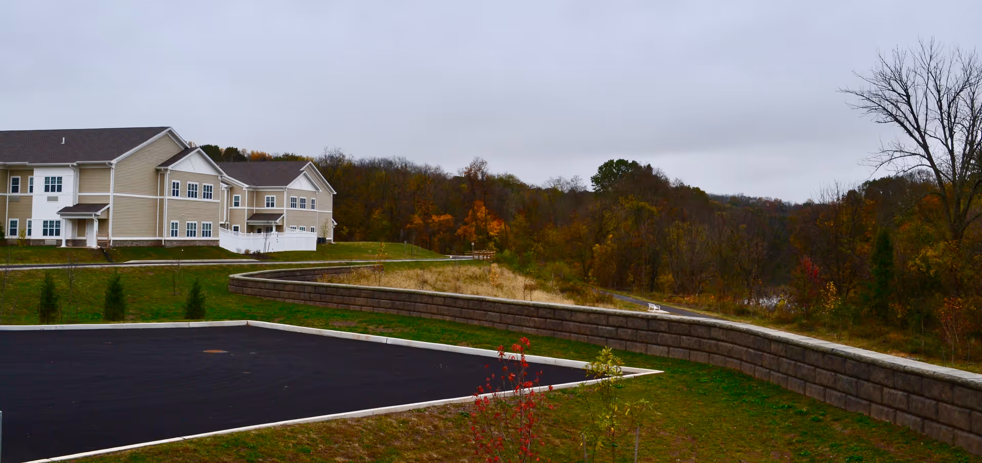 Exterior view of a beige two-story senior living facility building with white trim, surrounded by green grass, a paved parking lot, and a retaining wall. Trees with autumn foliage are visible in the background under a cloudy sky.