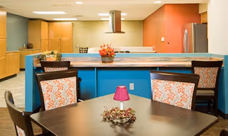 Interior view of a communal dining and kitchen area with a dark wooden table and four chairs featuring floral upholstery. A small purple lamp and decorative wreath are on the table. The kitchen area has wooden cabinets, a stainless steel refrigerator, and a blue counter with a flower arrangement on it. The walls are painted in shades of beige, blue, and red.