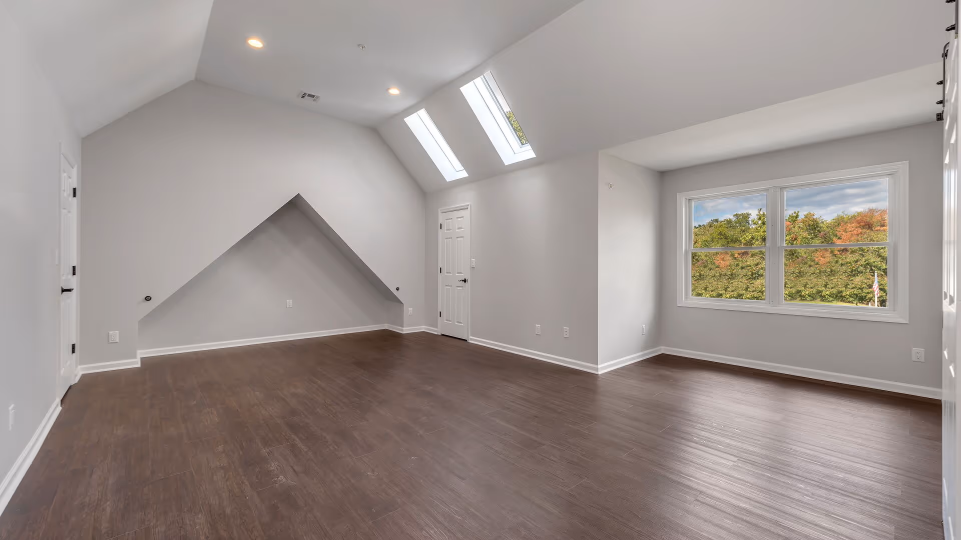 Empty room with wooden flooring, light gray walls, a door, two skylights on a slanted ceiling, and a large window showing green trees outside.