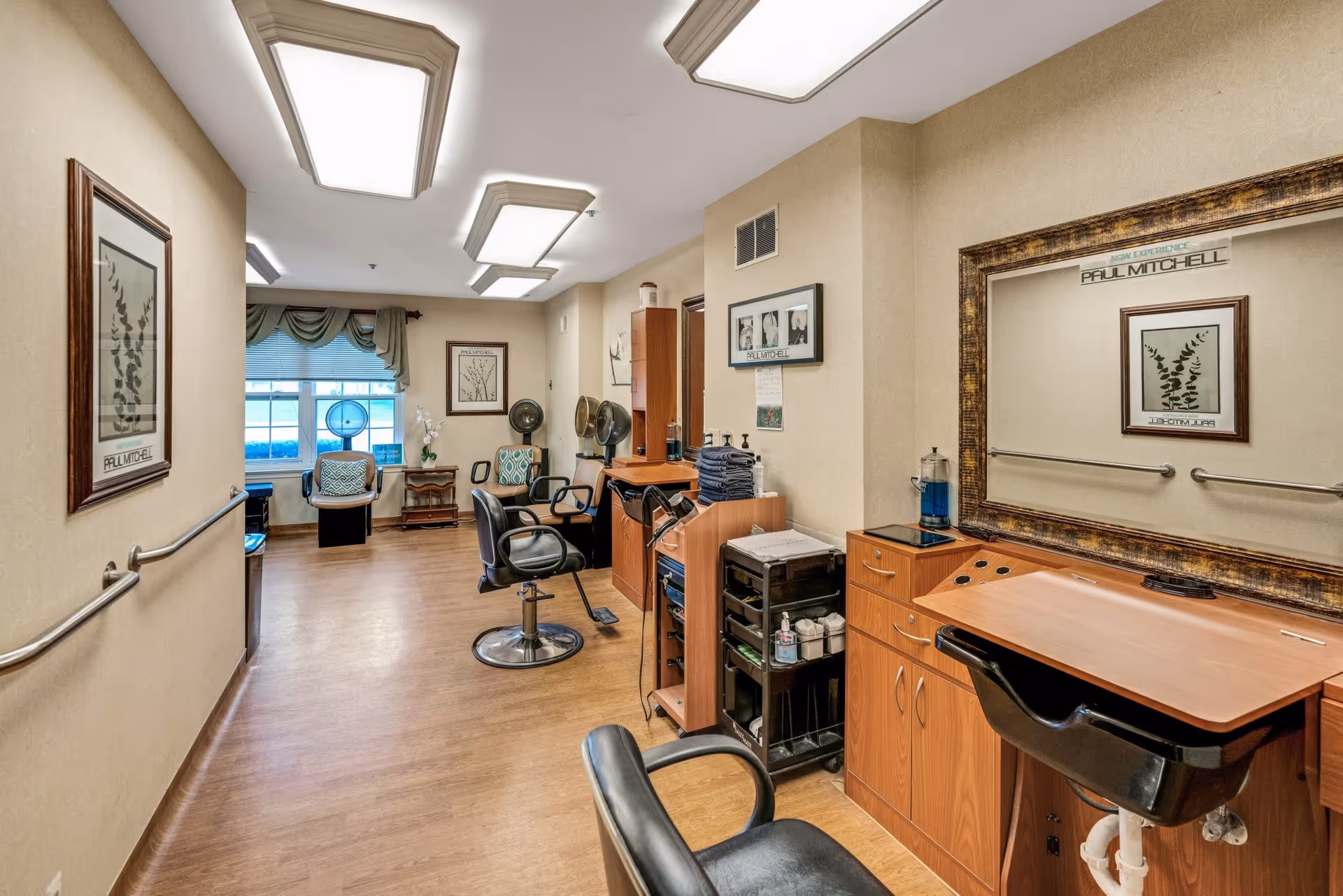 Interior view of a hair salon area in a senior living facility with wooden flooring, salon chairs, mirrors, hair dryers, and framed artwork on the walls. There is a window with green curtains and a seating area with two chairs and a small table near the window.