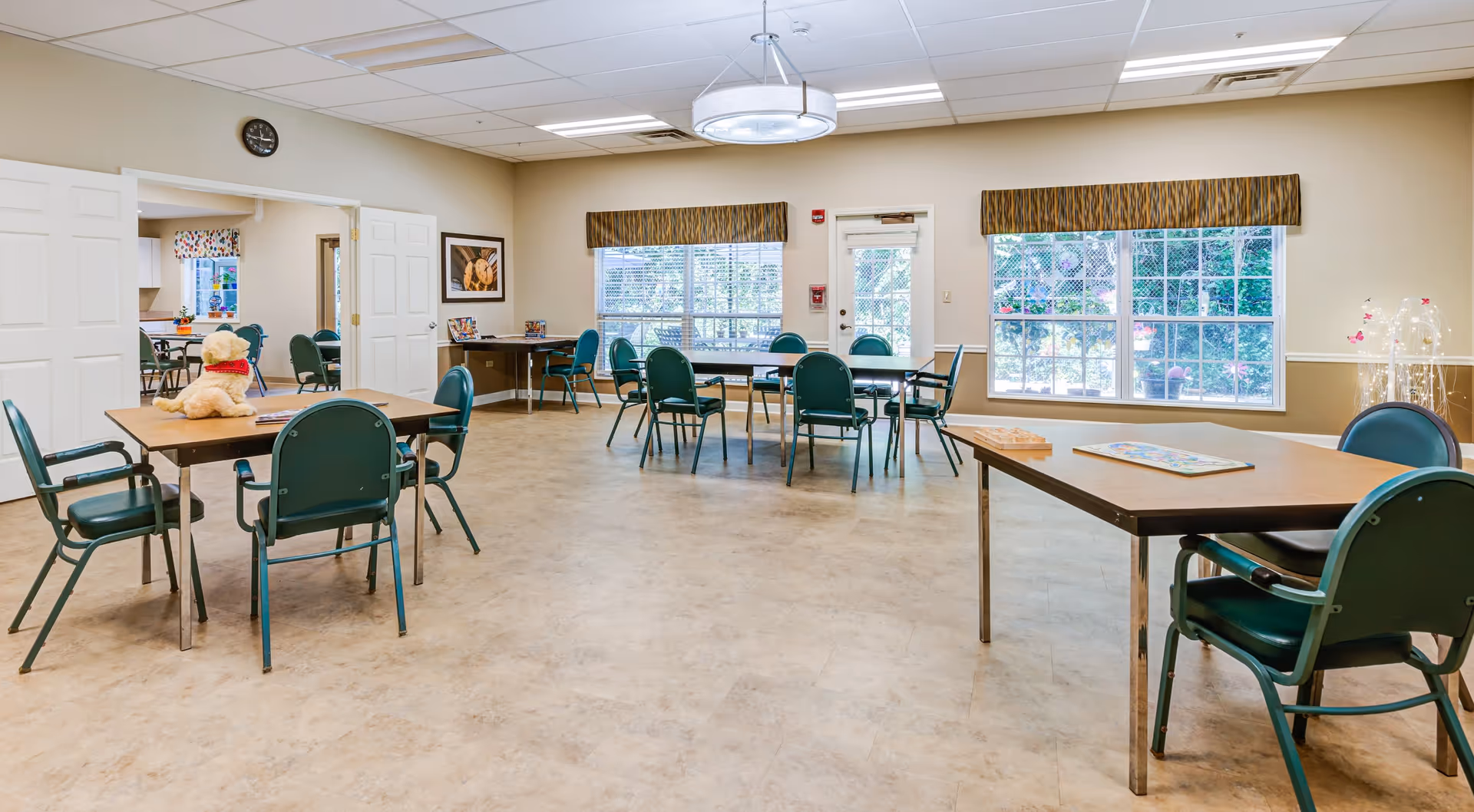 A spacious, well-lit common room with several tables and green chairs arranged around the room. There are large windows with patterned valances letting in natural light, a door leading outside, and a clock on the wall. One table has a stuffed dog toy on it, and another table has a board game. The room has a clean, welcoming atmosphere with beige walls and a light-colored floor.