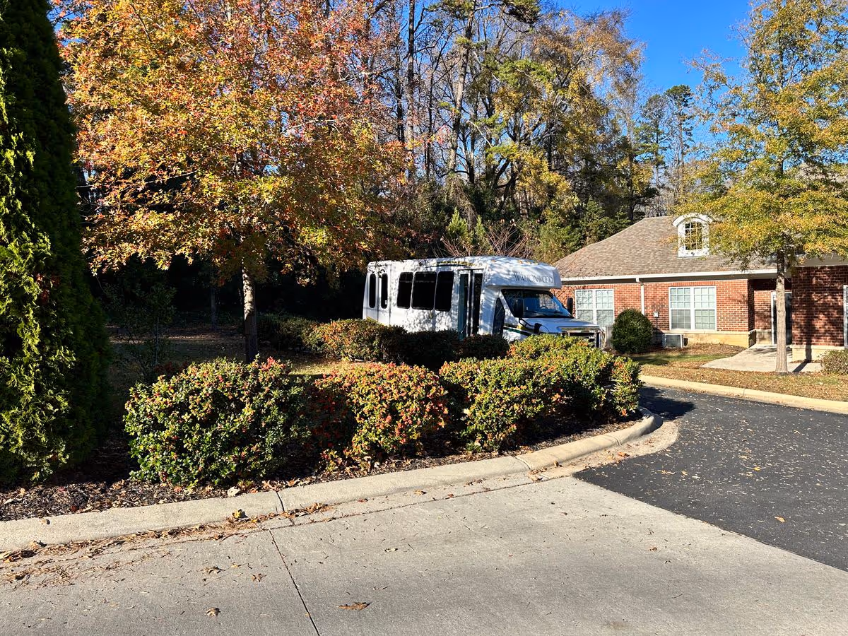 A white shuttle bus parked behind hedges in front of a brick building with trees and autumn foliage.