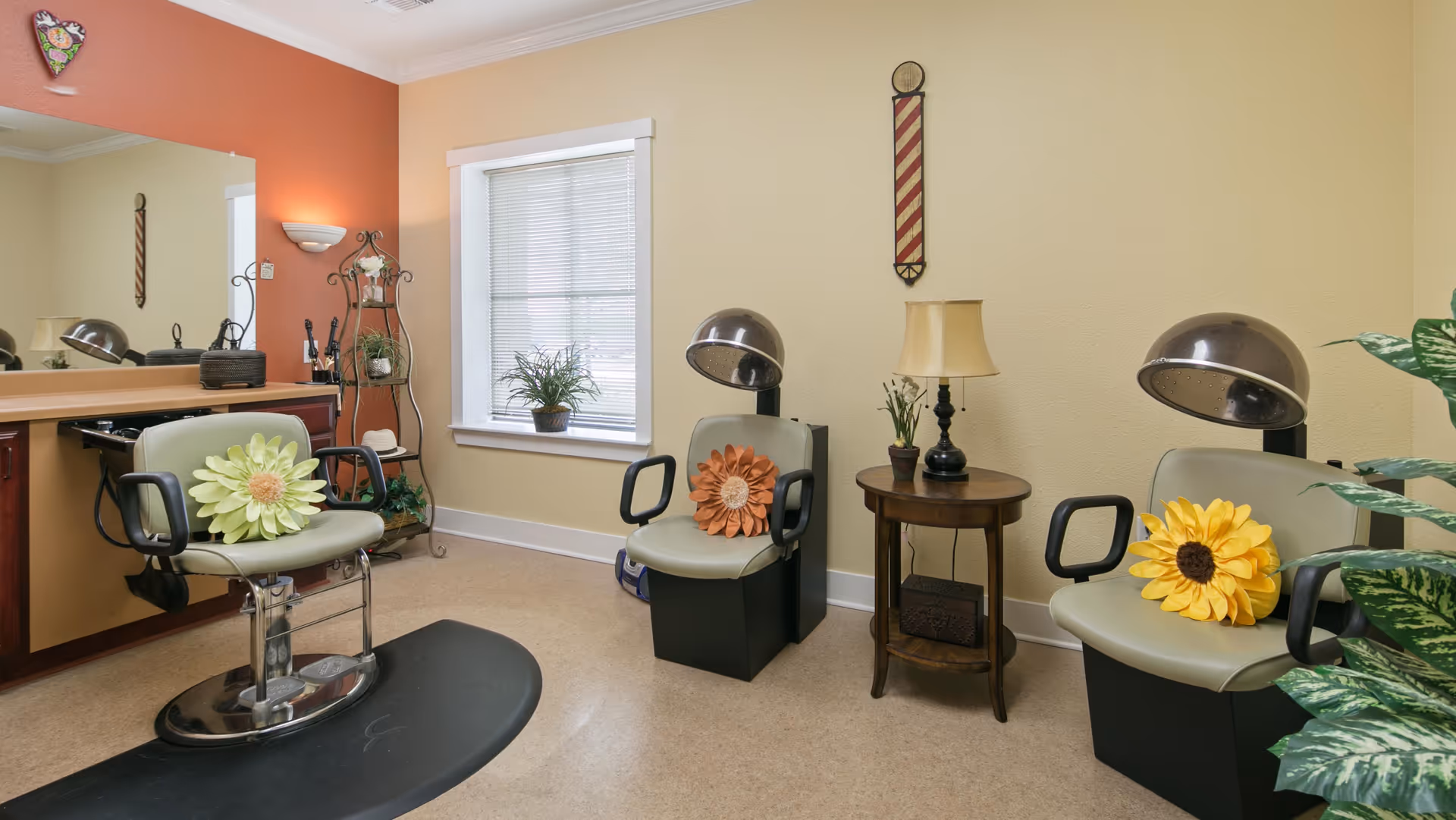 Interior view of a salon area in an assisted living facility with three salon chairs, two equipped with hair dryers. Each chair has a large decorative flower pillow. There is a window with blinds, a small round wooden table with a lamp and plants, and a large mirror on the left wall. The walls are painted in warm tones with a decorative wall hanging and some plants visible.