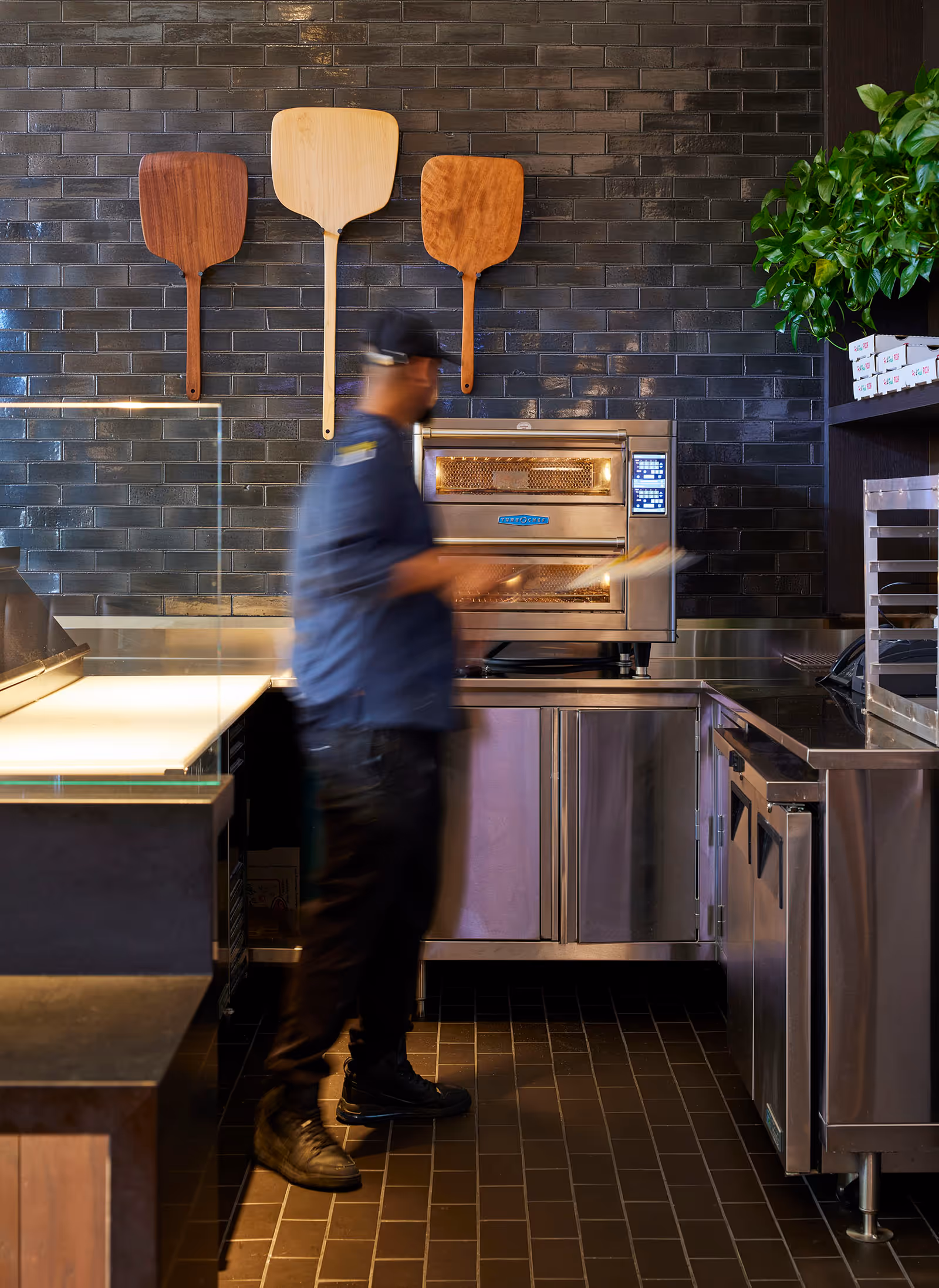 A person in a blue shirt and black cap is standing in a kitchen area with a stainless steel oven and cabinets. Three wooden pizza peels are hanging on a dark tiled wall above the oven. There is a green plant on the right side and some stacked boxes on a shelf.