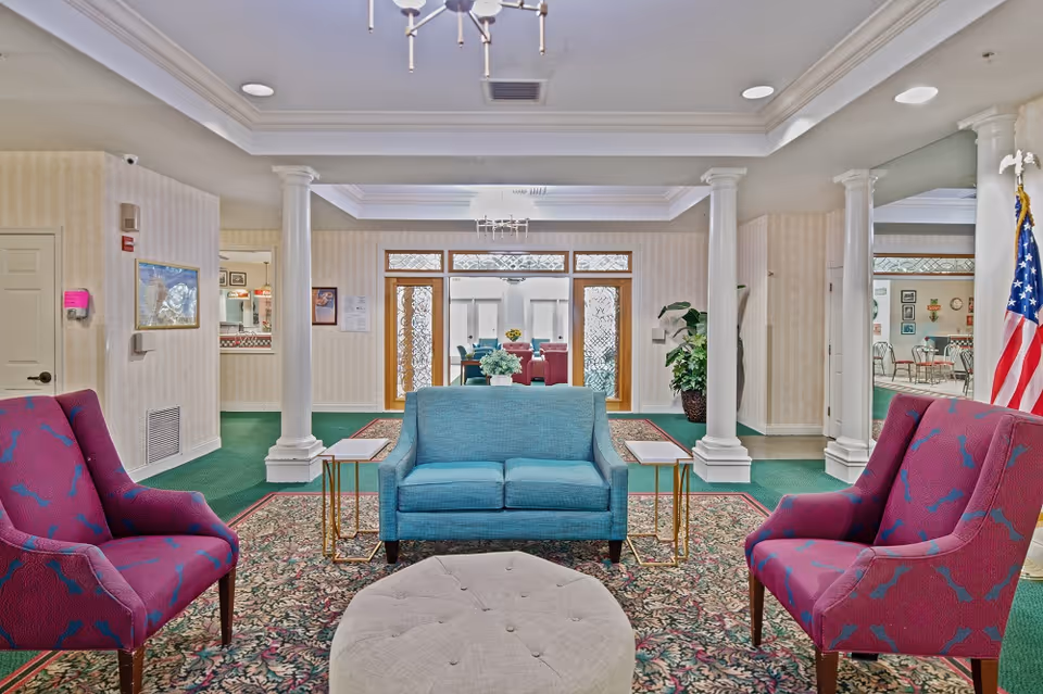 A seating area lobby with a blue loveseat, two purple armchairs, white columns and an American flag.