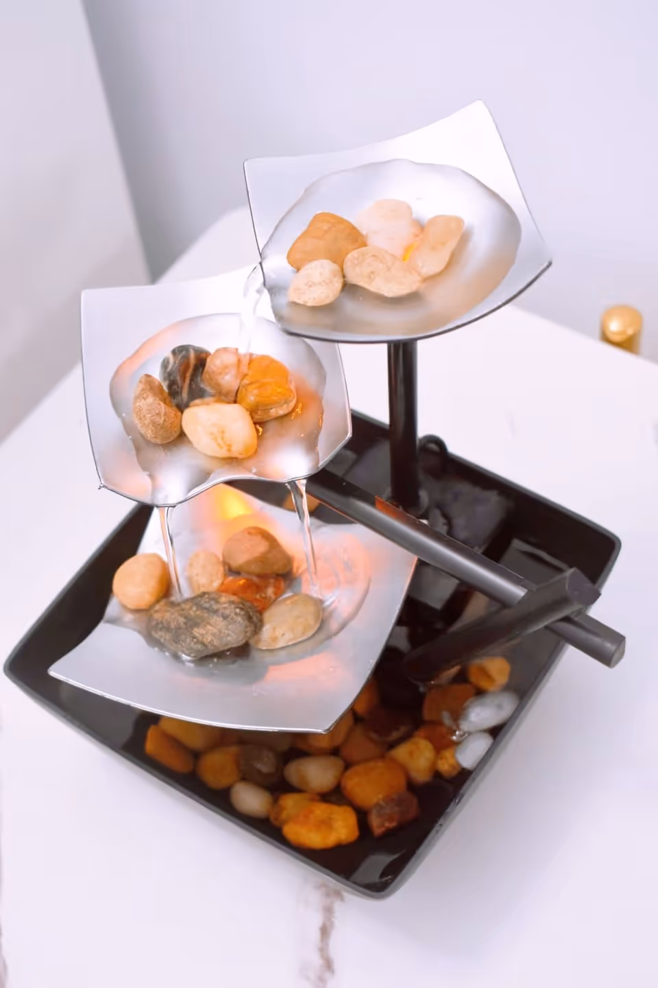 A small tabletop water fountain with three metal leaf-shaped tiers, each holding smooth decorative stones. Water flows gently from the top tier to the bottom, and the fountain is placed on a white surface.